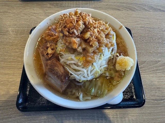 A white bowl of ramen on a black tray atop a wooden table, filled with broth, noodles, bean sprouts, cabbage, a thick slice of braised pork, minced garlic, and topped with crispy fried onions or garlic chips.