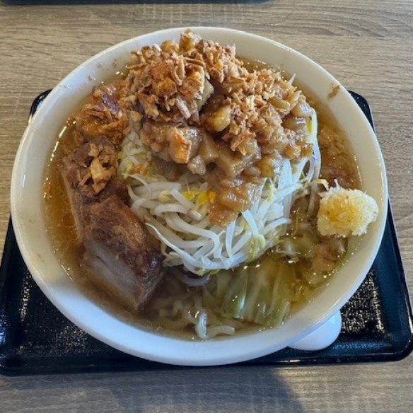 A white bowl of ramen on a black tray atop a wooden table, filled with broth, noodles, bean sprouts, cabbage, a thick slice of braised pork, minced garlic, and topped with crispy fried onions or garlic chips.